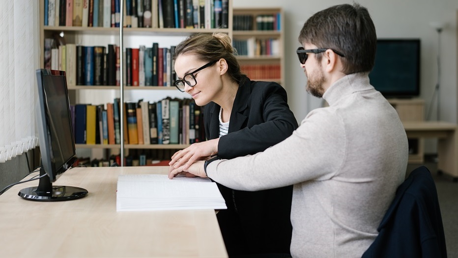 Eine Frau hilft einem blinden Mann beim Blindenschrift lesen.