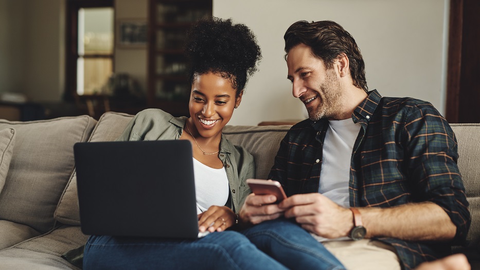 The picture shows a man and a woman casually sitting on a sofa and smiling while looking at the screen of a laptop on the woman`s lap while the man is holding a smartphone.